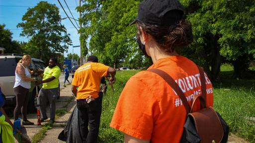 Duquesne Light workers are seen picking up trash in the background with a woman in the foreground wearing an orange DLC shirt that says "Volunteer" on the back